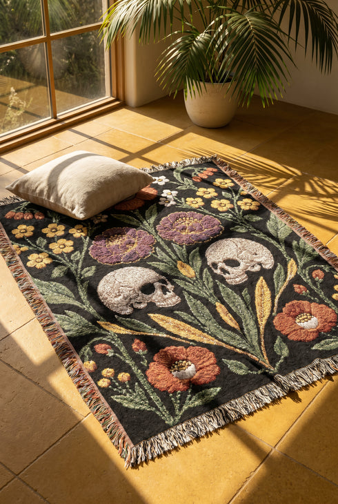 Woven cotton throw blanket with fringed edges and a black ground. Two cream skulls sit side by side at the center of a symmetrical folk garden. A terracotta poppy crowns the top, plum peonies bloom in the middle, and clusters of golden-yellow and white wildflowers, green leaves, and wheat-like sprigs fill the field, with more terracotta poppies along the bottom. The dimensional pattern is woven through a jacquard process, and the reverse shows a mirrored, softer version of the design.