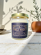 A Milk Moon candle in a clear glass jar with a gold lid. The ornate dark blue label has a celestial moon and star design. The hand-poured coconut apricot wax is a creamy off-white color, presented in a bright, clean setting to evoke a luxury spa feel.