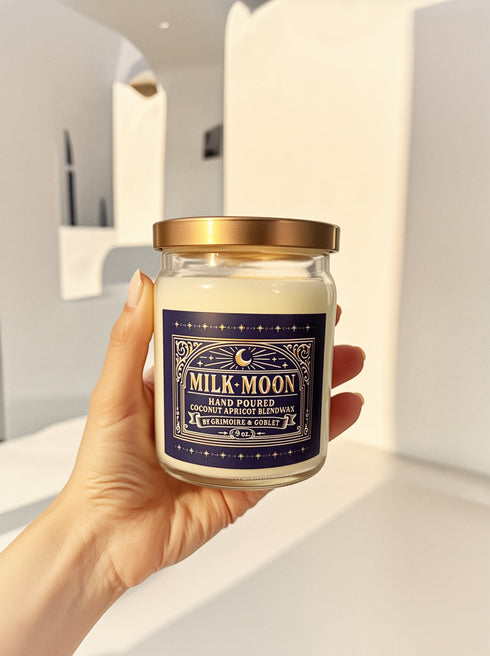 A Milk Moon candle in a clear glass jar with a gold lid. The ornate dark blue label has a celestial moon and star design. The hand-poured coconut apricot wax is a creamy off-white color, presented in a bright, clean setting to evoke a luxury spa feel.