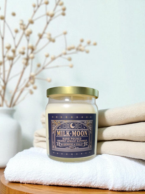 A Milk Moon candle in a clear glass jar with a gold lid. The ornate dark blue label has a celestial moon and star design. The hand-poured coconut apricot wax is a creamy off-white color, presented in a bright, clean setting to evoke a luxury spa feel.