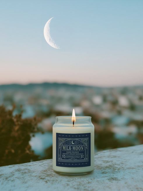 A Milk Moon candle in a clear glass jar with a gold lid. The ornate dark blue label has a celestial moon and star design. The hand-poured coconut apricot wax is a creamy off-white color, presented in a bright, clean setting to evoke a luxury spa feel.