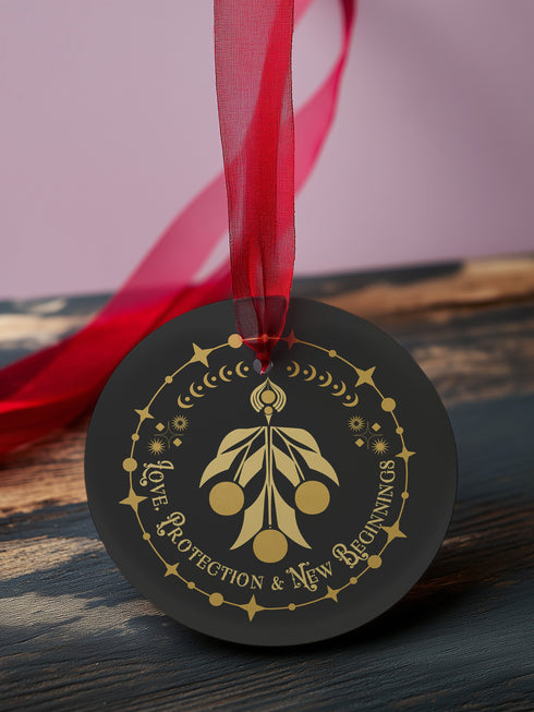 Round metal ornament featuring golden mistletoe design with Art Deco border incorporating stars and moon phases. 3.5-inch diameter piece with "Love, Protection & New Beginnings" text and crimson ribbon. Double-sided design on black background.
