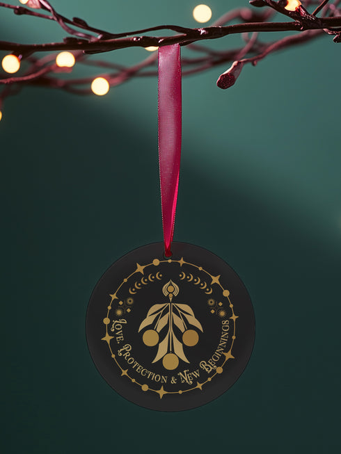 Round metal ornament featuring golden mistletoe design with Art Deco border incorporating stars and moon phases. 3.5-inch diameter piece with "Love, Protection & New Beginnings" text and crimson ribbon. Double-sided design on black background.