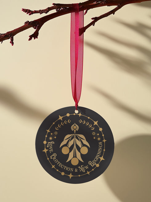 Round metal ornament featuring golden mistletoe design with Art Deco border incorporating stars and moon phases. 3.5-inch diameter piece with "Love, Protection & New Beginnings" text and crimson ribbon. Double-sided design on black background.