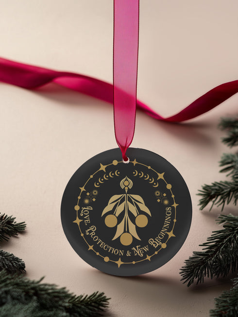 Round metal ornament featuring golden mistletoe design with Art Deco border incorporating stars and moon phases. 3.5-inch diameter piece with "Love, Protection & New Beginnings" text and crimson ribbon. Double-sided design on black background.