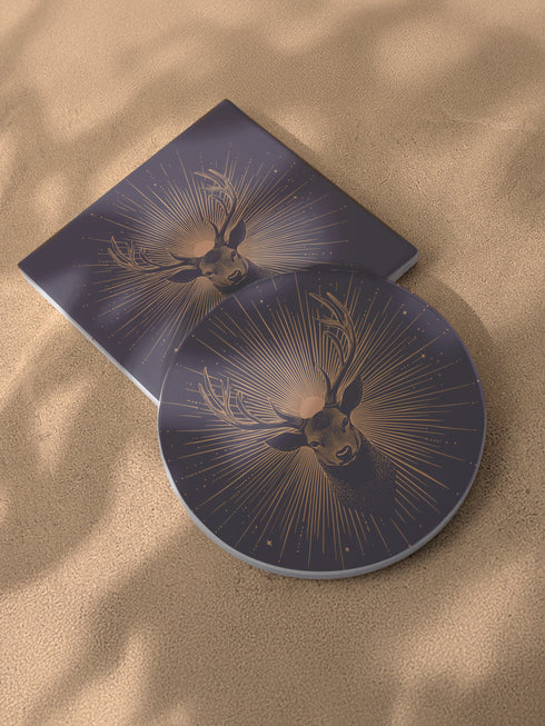 Ceramic coasters in round and square shapes featuring a detailed deer head with branching antlers on a deep navy background. Metallic gold sunburst rays radiate from behind the deer, with scattered stars throughout the design. Each coaster has a protective cork backing.