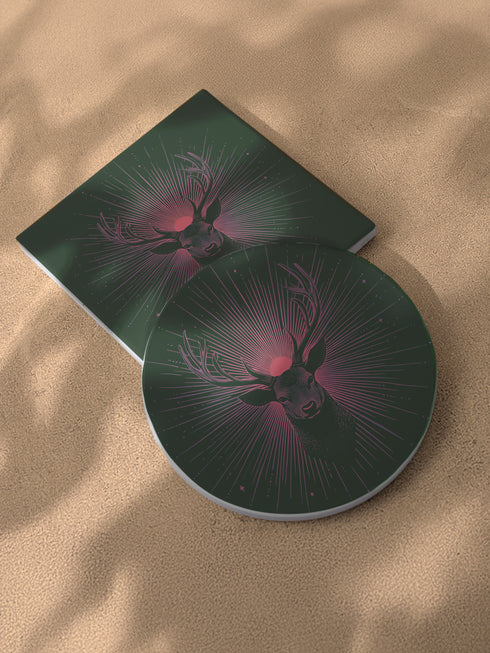 Ceramic coasters in round and square shapes featuring a detailed deer head with majestic antlers against a deep forest green background. A pink-tinged radiant sunburst pattern emanates from behind the deer's head, with delicate stars scattered throughout the design. Cork backing protection.