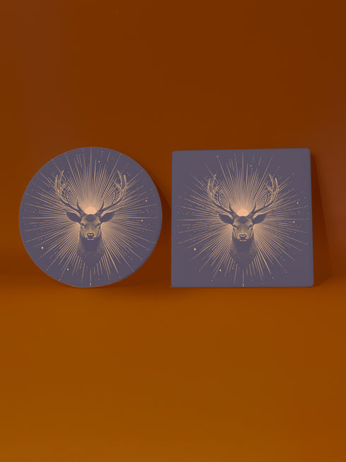 Ceramic coasters in round and square shapes featuring a detailed deer head with branching antlers on a deep navy background. Metallic gold sunburst rays radiate from behind the deer, with scattered stars throughout the design. Each coaster has a protective cork backing.