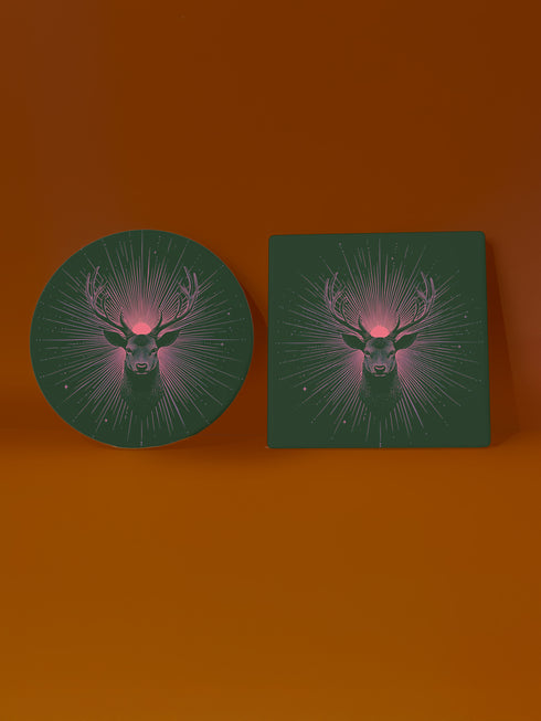 Ceramic coasters in round and square shapes featuring a detailed deer head with majestic antlers against a deep forest green background. A pink-tinged radiant sunburst pattern emanates from behind the deer's head, with delicate stars scattered throughout the design. Cork backing protection.