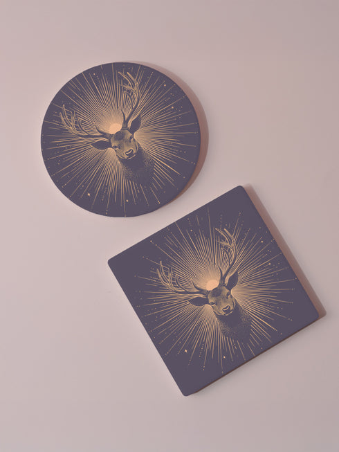 Ceramic coasters in round and square shapes featuring a detailed deer head with branching antlers on a deep navy background. Metallic gold sunburst rays radiate from behind the deer, with scattered stars throughout the design. Each coaster has a protective cork backing.