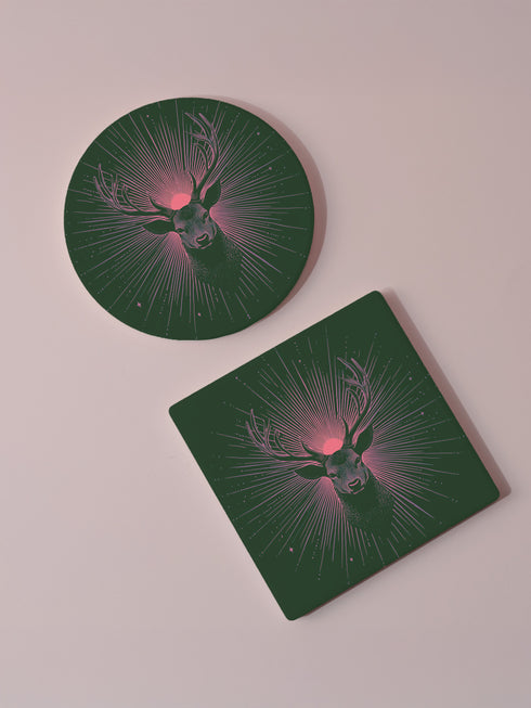 Ceramic coasters in round and square shapes featuring a detailed deer head with majestic antlers against a deep forest green background. A pink-tinged radiant sunburst pattern emanates from behind the deer's head, with delicate stars scattered throughout the design. Cork backing protection.