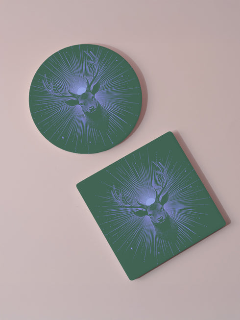 Ceramic coasters in round and square shapes featuring a detailed deer head with antlers on a forest green background, surrounded by radiating periwinkle blue sunburst lines and small stars, with protective cork backing.