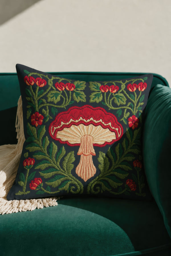 Square decorative pillow cover with a deep midnight navy ground and a symmetrical Art Nouveau botanical design rendered in a faux embroidery style. A large central mushroom anchors the composition, its scarlet ruffled cap fanning over cream and ivory gills above a pale peach stem. Curling green ferns, leafy vines, crimson flower buds, and small red blossoms frame the toadstool on every side. The printed satin-stitch detailing mimics hand-stitched thread across the entire face of the cover.