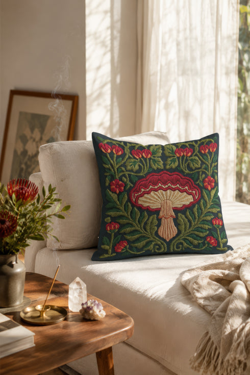 Square decorative pillow cover with a deep midnight navy ground and a symmetrical Art Nouveau botanical design rendered in a faux embroidery style. A large central mushroom anchors the composition, its scarlet ruffled cap fanning over cream and ivory gills above a pale peach stem. Curling green ferns, leafy vines, crimson flower buds, and small red blossoms frame the toadstool on every side. The printed satin-stitch detailing mimics hand-stitched thread across the entire face of the cover.