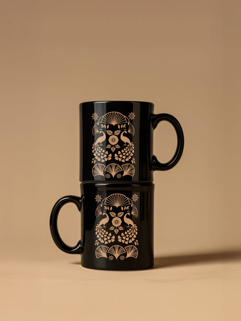A black ceramic mug featuring an Art Deco-inspired design in rose gold. The symmetrical pattern shows two peacocks with fan-shaped tails, surrounded by ginkgo fan motifs and floral elements. The central design includes a stylized flower with leaves, and decorative stars accent the composition. The mug has a classic C-shaped handle.