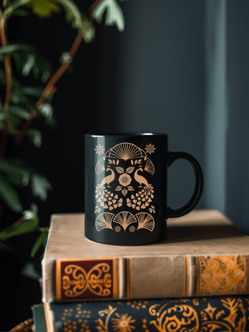 A black ceramic mug featuring an Art Deco-inspired design in rose gold. The symmetrical pattern shows two peacocks with fan-shaped tails, surrounded by ginkgo fan motifs and floral elements. The central design includes a stylized flower with leaves, and decorative stars accent the composition. The mug has a classic C-shaped handle.
