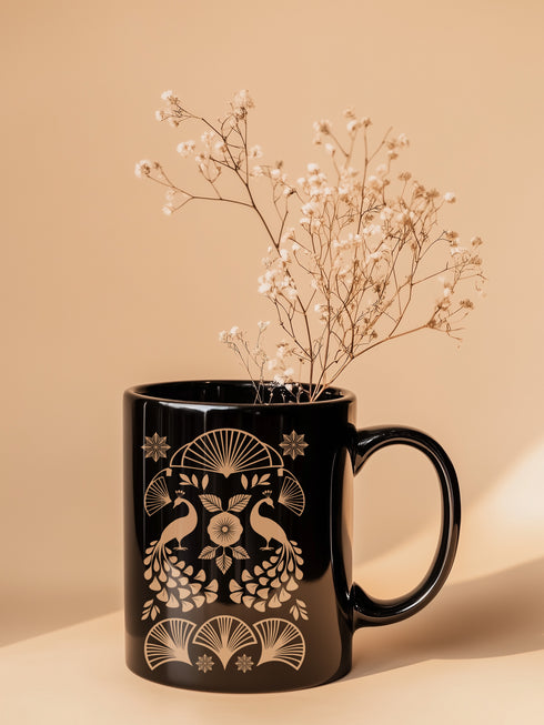 A black ceramic mug featuring an Art Deco-inspired design in rose gold. The symmetrical pattern shows two peacocks with fan-shaped tails, surrounded by ginkgo fan motifs and floral elements. The central design includes a stylized flower with leaves, and decorative stars accent the composition. The mug has a classic C-shaped handle.