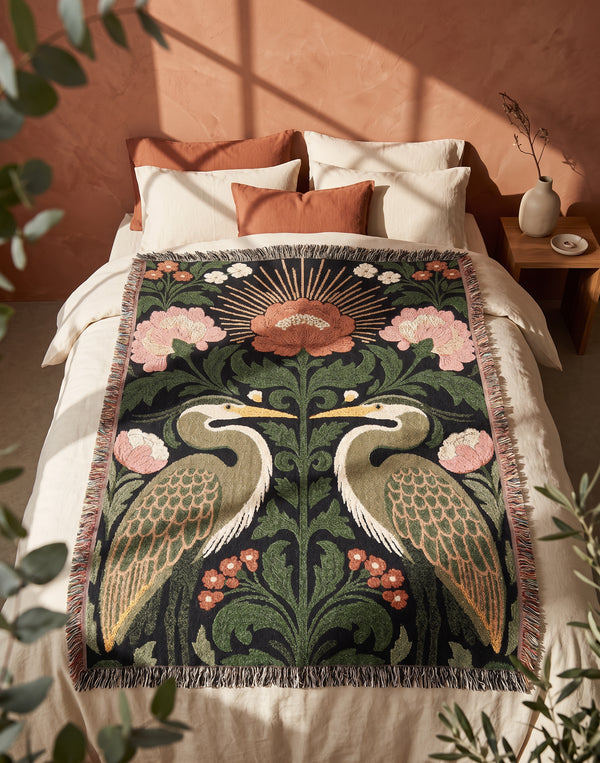 Jacquard woven cotton throw blanket, woven rather than printed, shown flat. Two mirrored great blue herons flank a central radial sunburst, surrounded by botanical blooms and scrollwork. The charcoal black ground carries antique gold ochre sunbeams, blush pink blossoms, deep forest green leaves, muted rust berry clusters, and cream toned herons. A lush multi-colored fringe of black, ochre, and sage threads edges the blanket, and the reverse shows an inverted charcoal grey version of the same design.