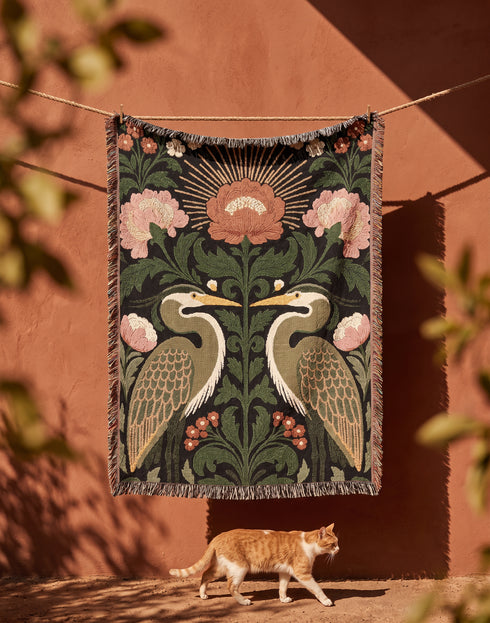 Jacquard woven cotton throw blanket, woven rather than printed, shown flat. Two mirrored great blue herons flank a central radial sunburst, surrounded by botanical blooms and scrollwork. The charcoal black ground carries antique gold ochre sunbeams, blush pink blossoms, deep forest green leaves, muted rust berry clusters, and cream toned herons. A lush multi-colored fringe of black, ochre, and sage threads edges the blanket, and the reverse shows an inverted charcoal grey version of the same design.
