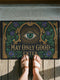 Rectangular doormat printed on a deep black ground with a faux embroidery effect that imitates antique crewelwork. A large green eye sits at the center within an antique gold radial sunburst, with ornate gold serif lettering below reading MAY ONLY GOOD ENTER. Green acanthus leaf scrollwork and antique gold architectural columns run along the borders, accented with small five petaled purple flowers. A sleek black trim frames all four edges of the flat printed surface.