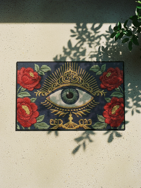 A rectangular navy blue Grimoire and Goblet doormat featuring a large central evil eye motif with a green iris and antique gold radiating lashes. The eye is surrounded by ornate gold filigree and four red peonies with green leaves in the corners. The design utilizes a high-quality 2D print technique to mimic the visual texture of vintage needlepoint embroidery, creating a trompe-l'œil effect on a flat 100% polyester surface.