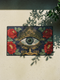 A rectangular navy blue Grimoire and Goblet doormat featuring a large central evil eye motif with a green iris and antique gold radiating lashes. The eye is surrounded by ornate gold filigree and four red peonies with green leaves in the corners. The design utilizes a high-quality 2D print technique to mimic the visual texture of vintage needlepoint embroidery, creating a trompe-l'œil effect on a flat 100% polyester surface.