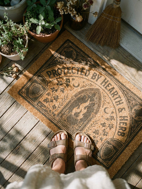 Rectangular coir doormat woven from natural brown coconut fiber with a bristly low pile. A bold black printed design fills the surface: a pointed archway frames a cast iron cauldron with rising flames at the center, flanked by winding ivy vines and small crescent moons. Arched lettering across the top reads PROTECTED BY HEARTH AND HERB. The four corners hold medieval shields and ornamental medallions, all enclosed by a thick black rectangular border with finished edges.