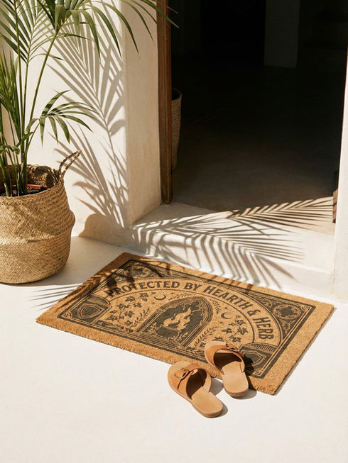 Rectangular coir doormat woven from natural brown coconut fiber with a bristly low pile. A bold black printed design fills the surface: a pointed archway frames a cast iron cauldron with rising flames at the center, flanked by winding ivy vines and small crescent moons. Arched lettering across the top reads PROTECTED BY HEARTH AND HERB. The four corners hold medieval shields and ornamental medallions, all enclosed by a thick black rectangular border with finished edges.