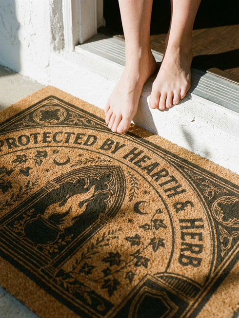 Rectangular coir doormat woven from natural brown coconut fiber with a bristly low pile. A bold black printed design fills the surface: a pointed archway frames a cast iron cauldron with rising flames at the center, flanked by winding ivy vines and small crescent moons. Arched lettering across the top reads PROTECTED BY HEARTH AND HERB. The four corners hold medieval shields and ornamental medallions, all enclosed by a thick black rectangular border with finished edges.