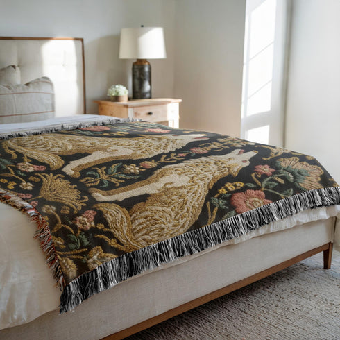 A rectangular, 100% woven cotton throw blanket with a detailed jacquard design. The throw features a symmetrical, vertical pattern of two howling wolves in profile, rendered in shades of beige, gold, and brown against a solid black background. The wolves are surrounded by intricate floral and foliage motifs. The thick, textured weave is visible. The blanket is finished on all four sides with a two-inch, multi-colored fringe that includes black, white, red, and gold threads.