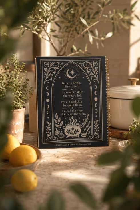 Spiral bound hardcover recipe journal lying flat. The matte black front cover is illustrated with light beige botanical vines, small crescent moons, a radial starburst, and a central cauldron releasing curls of steam, with classical lettering reading Spells of Bone and Broth. A black metal wire-o coil binds the left edge. The interior shows bright white pages printed with a structured recipe template, including labeled sections for ingredients and instructions, over a rigid chipboard backing.