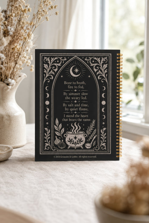 Spiral bound hardcover recipe journal lying flat. The matte black front cover is illustrated with light beige botanical vines, small crescent moons, a radial starburst, and a central cauldron releasing curls of steam, with classical lettering reading Spells of Bone and Broth. A black metal wire-o coil binds the left edge. The interior shows bright white pages printed with a structured recipe template, including labeled sections for ingredients and instructions, over a rigid chipboard backing.
