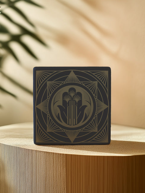 A black and gold ceramic coaster featuring an intricate Hamsa Hand design with celestial and art deco influences.