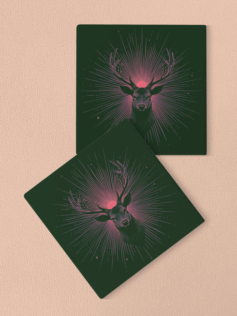 Ceramic coasters in round and square shapes featuring a detailed deer head with majestic antlers against a deep forest green background. A pink-tinged radiant sunburst pattern emanates from behind the deer's head, with delicate stars scattered throughout the design. Cork backing protection.