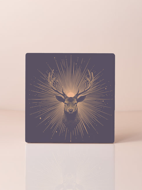 Ceramic coasters in round and square shapes featuring a detailed deer head with branching antlers on a deep navy background. Metallic gold sunburst rays radiate from behind the deer, with scattered stars throughout the design. Each coaster has a protective cork backing.