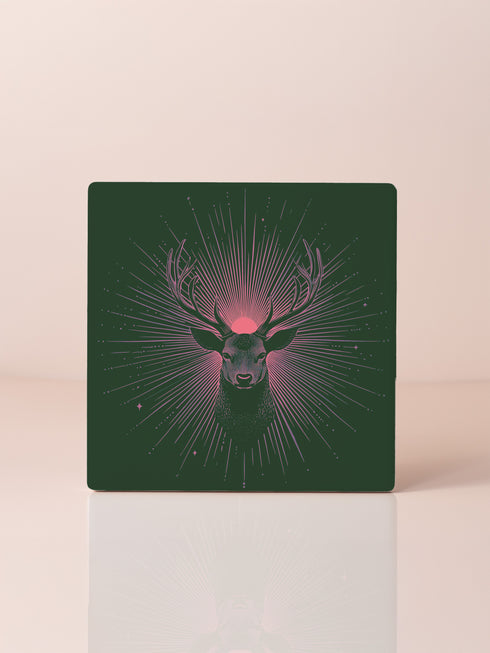 Ceramic coasters in round and square shapes featuring a detailed deer head with majestic antlers against a deep forest green background. A pink-tinged radiant sunburst pattern emanates from behind the deer's head, with delicate stars scattered throughout the design. Cork backing protection.