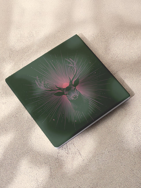 Ceramic coasters in round and square shapes featuring a detailed deer head with majestic antlers against a deep forest green background. A pink-tinged radiant sunburst pattern emanates from behind the deer's head, with delicate stars scattered throughout the design. Cork backing protection.