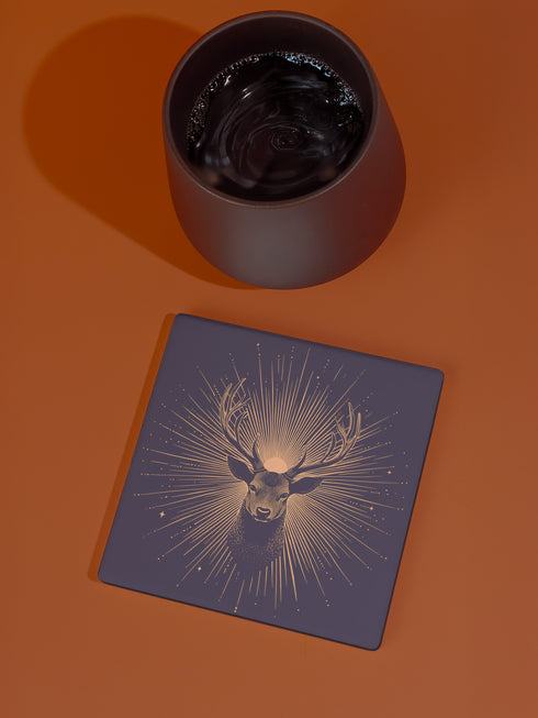 Ceramic coasters in round and square shapes featuring a detailed deer head with branching antlers on a deep navy background. Metallic gold sunburst rays radiate from behind the deer, with scattered stars throughout the design. Each coaster has a protective cork backing.