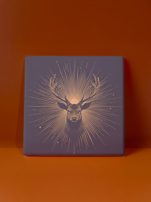 Ceramic coasters in round and square shapes featuring a detailed deer head with branching antlers on a deep navy background. Metallic gold sunburst rays radiate from behind the deer, with scattered stars throughout the design. Each coaster has a protective cork backing.