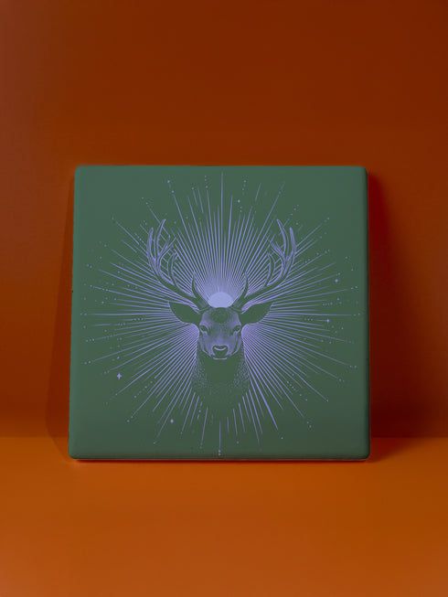 Ceramic coasters in round and square shapes featuring a detailed deer head with antlers on a forest green background, surrounded by radiating periwinkle blue sunburst lines and small stars, with protective cork backing.