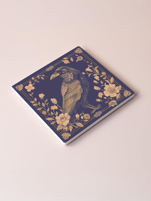 Square ceramic coaster featuring detailed illustration of a raven surrounded by golden florals and botanicals on navy background. Decorative border with dotted pattern frames the design.