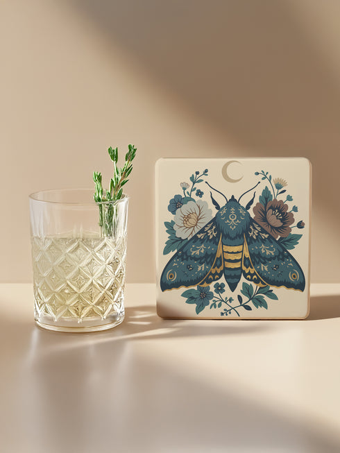 Square ceramic coaster, 4x4 inches, with cork backing. Cream tile features a luna-style moth with crescent moon and botanical florals in teal, blue, and gold. Smooth printed surface; clean, flat illustration. Celestial moth coaster for witchy, cottagecore décor and gifting. Square ceramic drink coaster with cork base, luna moth and moon design.