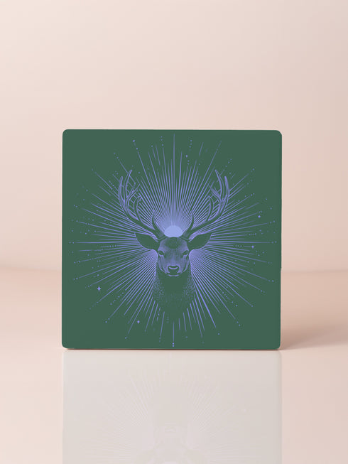 Ceramic coasters in round and square shapes featuring a detailed deer head with antlers on a forest green background, surrounded by radiating periwinkle blue sunburst lines and small stars, with protective cork backing.
