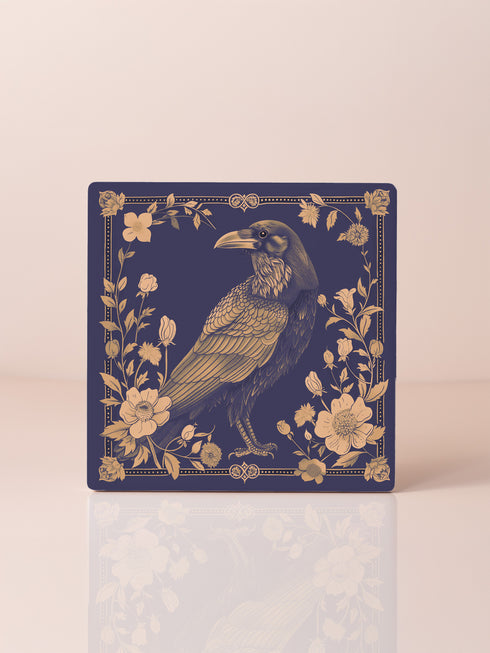 Square ceramic coaster featuring detailed illustration of a raven surrounded by golden florals and botanicals on navy background. Decorative border with dotted pattern frames the design.