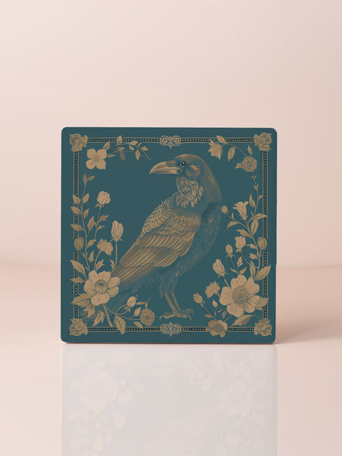 Square ceramic coaster featuring detailed gold raven illustration surrounded by botanical elements on emerald green background, with ornate border design and cork backing.