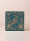 Square ceramic coaster featuring detailed gold raven illustration surrounded by botanical elements on emerald green background, with ornate border design and cork backing.