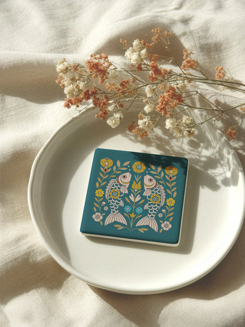 Square ceramic coaster, 4x4 inches, with cork backing. Teal tile features a folk-art twin fish motif framed by florals in mustard, cream, and soft blue. Smooth printed surface; crisp, flat color artwork. Witchy zodiac-inspired Pisces coaster for mystical home décor, coffee table protection, and gifting. Square ceramic drink coaster with cork base, teal fish design.