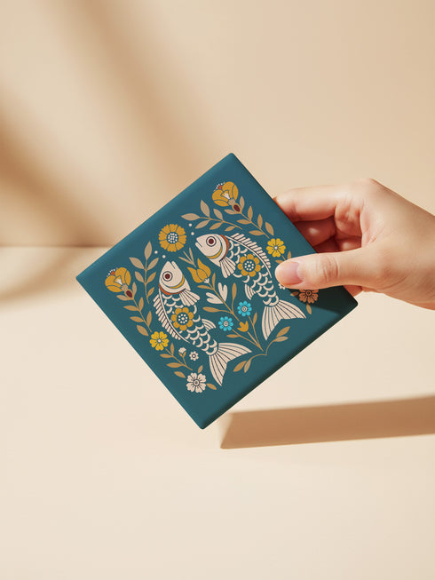 Square ceramic coaster, 4x4 inches, with cork backing. Teal tile features a folk-art twin fish motif framed by florals in mustard, cream, and soft blue. Smooth printed surface; crisp, flat color artwork. Witchy zodiac-inspired Pisces coaster for mystical home décor, coffee table protection, and gifting. Square ceramic drink coaster with cork base, teal fish design.