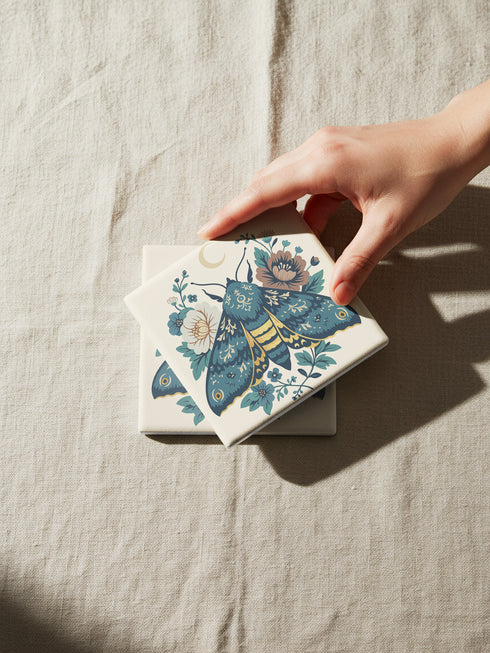 Square ceramic coaster, 4x4 inches, with cork backing. Cream tile features a luna-style moth with crescent moon and botanical florals in teal, blue, and gold. Smooth printed surface; clean, flat illustration. Celestial moth coaster for witchy, cottagecore décor and gifting. Square ceramic drink coaster with cork base, luna moth and moon design.