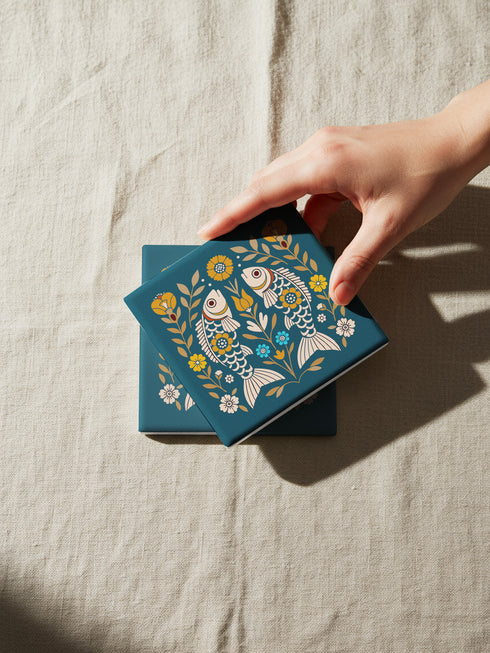 Square ceramic coaster, 4x4 inches, with cork backing. Teal tile features a folk-art twin fish motif framed by florals in mustard, cream, and soft blue. Smooth printed surface; crisp, flat color artwork. Witchy zodiac-inspired Pisces coaster for mystical home décor, coffee table protection, and gifting. Square ceramic drink coaster with cork base, teal fish design.